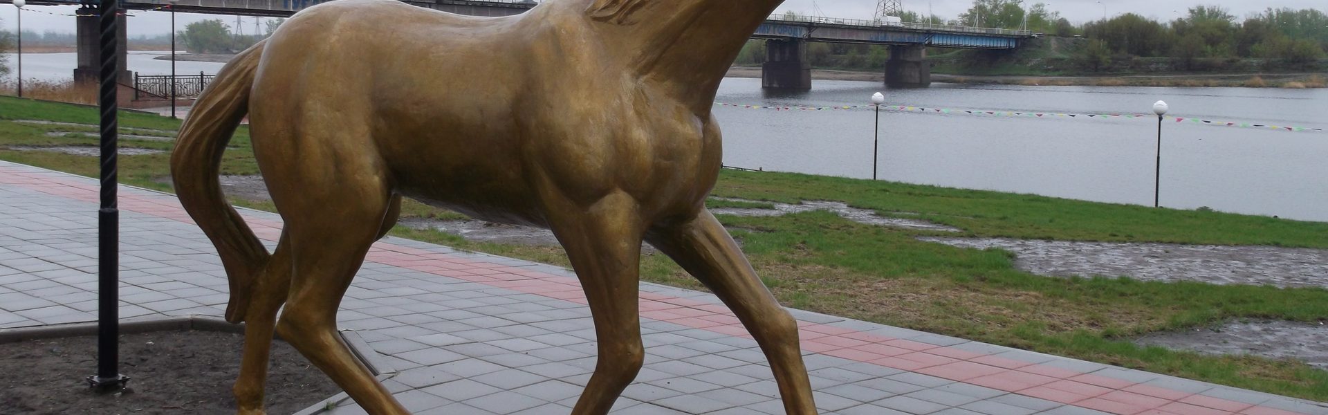 Набережная Тобола в Костанае весной — бронзовая статуя коня, мост через реку и флажки вдоль променада